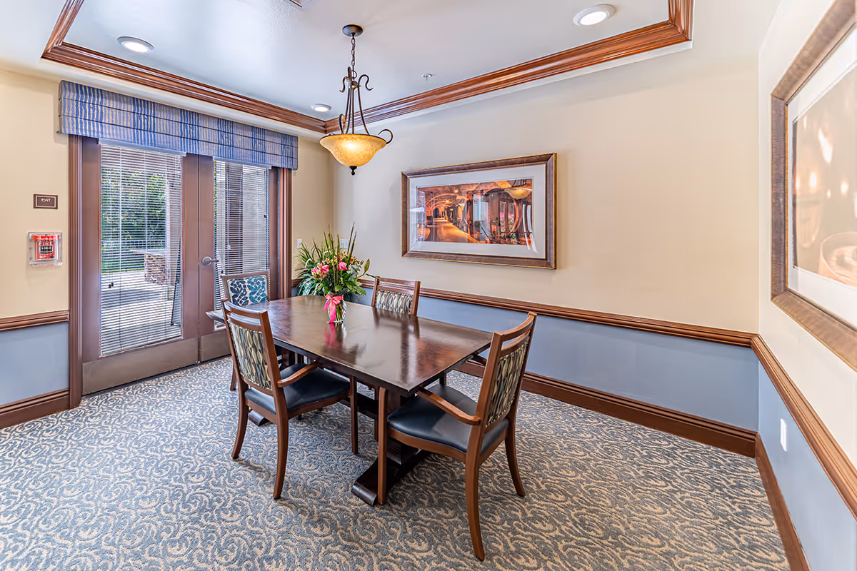A small dining room with a rectangular wooden table surrounded by four chairs with patterned upholstery. A vase with a floral arrangement sits on the table. The room has patterned carpet, beige walls with blue wainscoting, and crown molding. A framed picture hangs on the wall, and double glass doors with blinds lead outside. A ceiling light fixture hangs above the table.