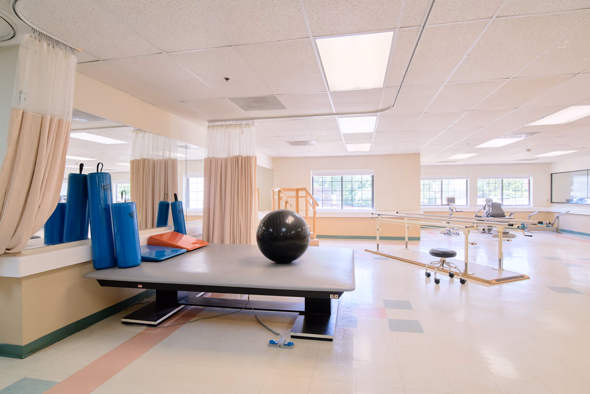 Bright rehabilitation/physical therapy room with a treatment table, large exercise ball, foam rollers, parallel bars and windows.