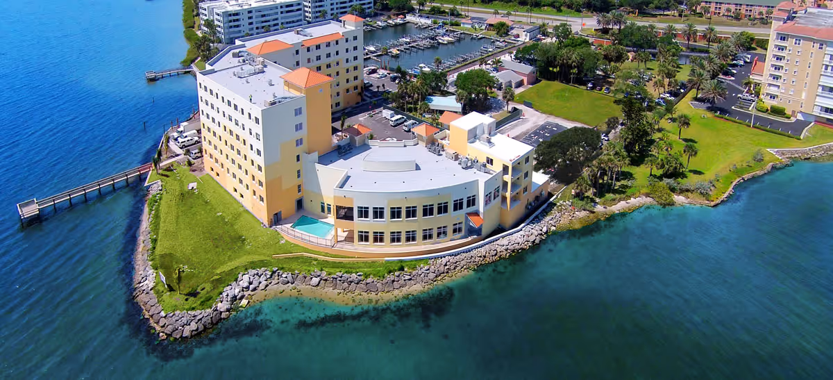 Aerial view of a waterfront senior living facility on a small peninsula with a pool, dock, marina, and surrounding green grounds.