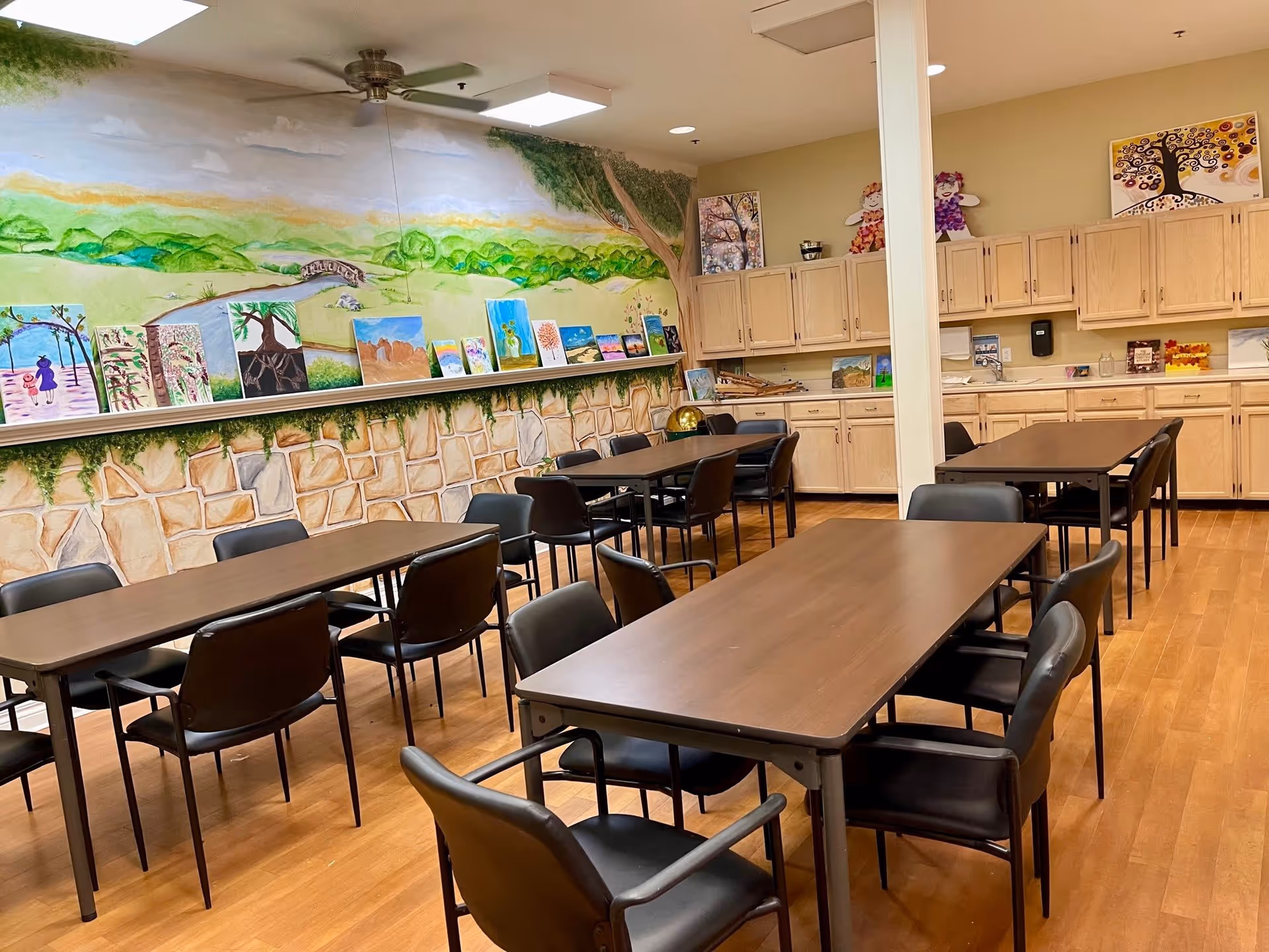 A room with several rectangular tables and black chairs arranged around them. The walls feature a mural of a scenic landscape with trees, hills, and a bridge, along with a display of various paintings on a ledge. Light wood cabinets line one wall, and the floor is a light wood laminate. The ceiling has a fan and recessed lighting.