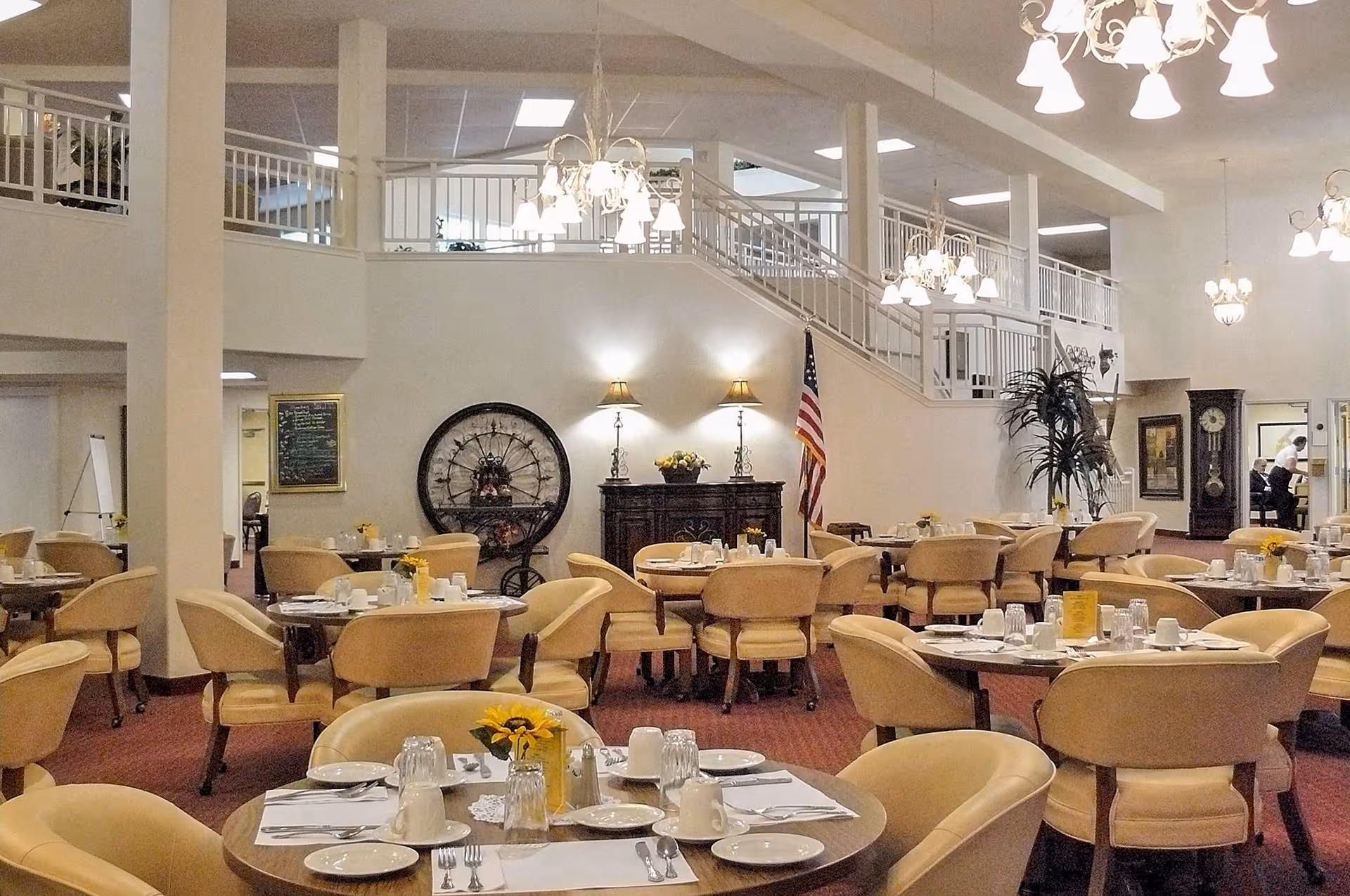 A spacious dining room with multiple round tables set with white plates, cups, silverware, and yellow flower centerpieces. The room features beige upholstered chairs, red carpet flooring, and several chandeliers hanging from the ceiling. There is a staircase with white railings leading to an upper level, an American flag, a large decorative clock, and a wooden cabinet with two lamps on it against the far wall.