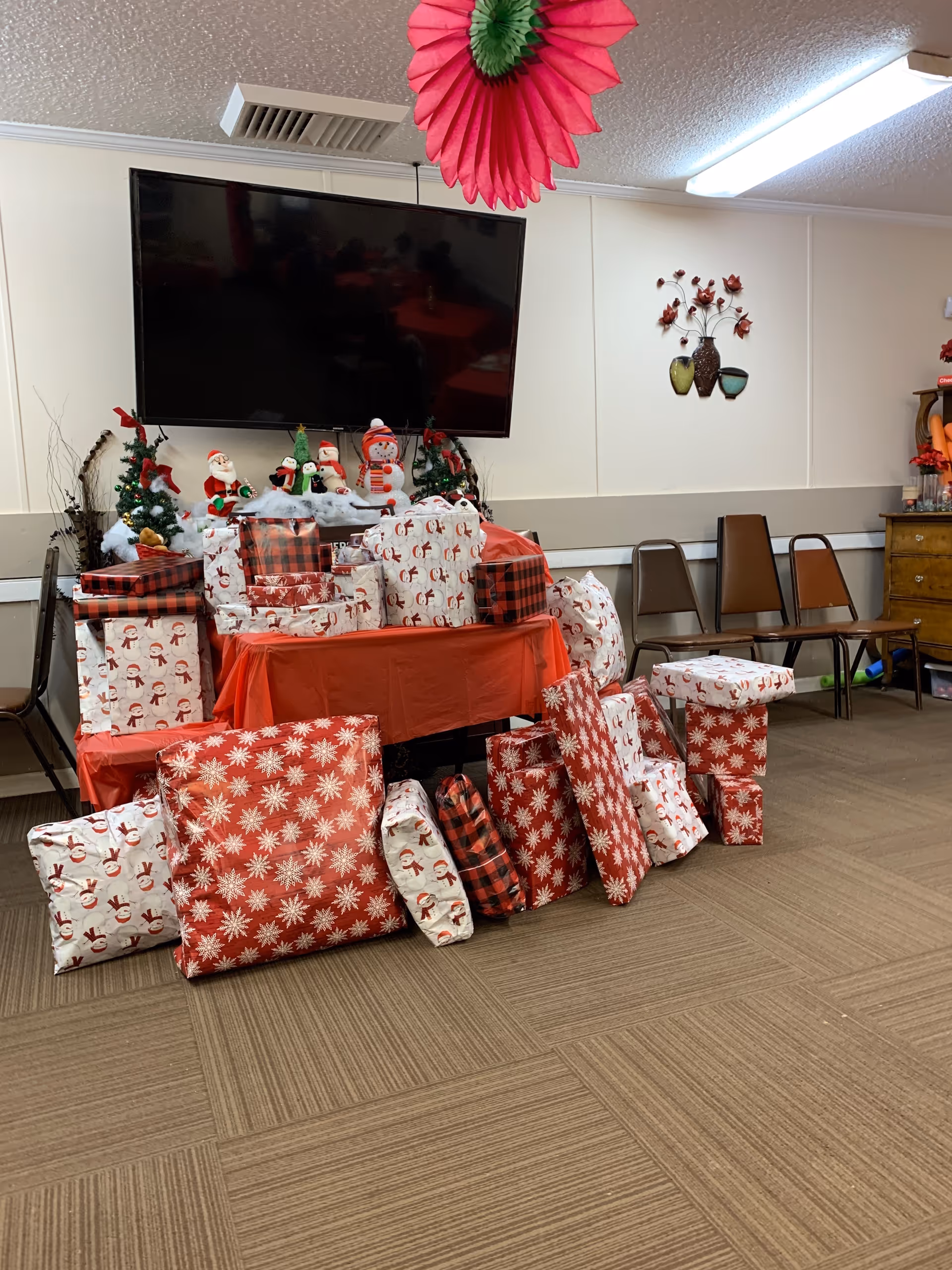 A room decorated for Christmas with a table covered in a red tablecloth and stacked with wrapped presents in festive paper. Behind the table are small Christmas decorations including Santa Claus figures, a snowman, and small Christmas trees. A large flat-screen TV is mounted on the wall above the table. Several brown chairs are lined up against the wall to the right, and a pink and green paper decoration hangs from the ceiling.