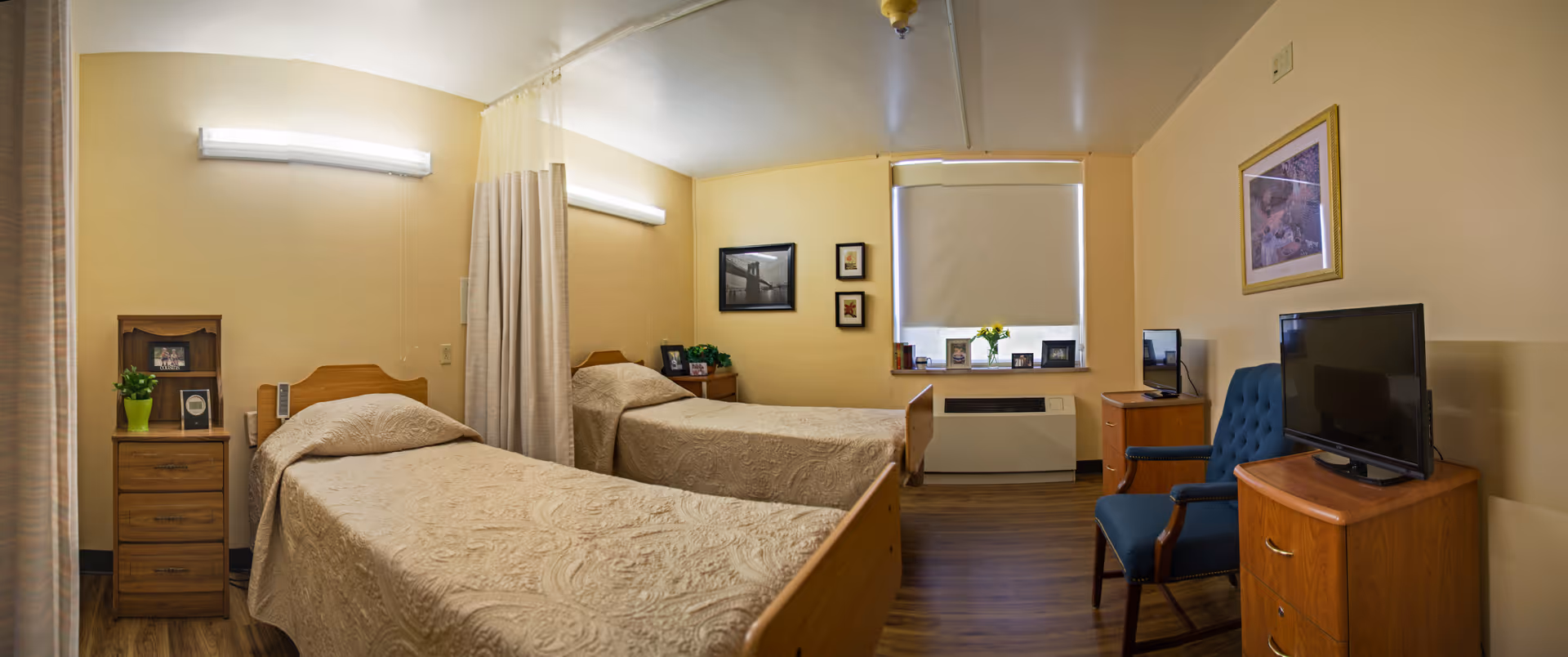 A rehabilitation facility room with two single beds separated by a privacy curtain. Each bed has a wooden headboard and beige patterned bedspread. There are wooden nightstands with framed photos and a small plant. The room has yellow walls, a window with a closed blind, a blue upholstered chair, a wooden dresser with a flat-screen TV, and framed pictures on the walls.