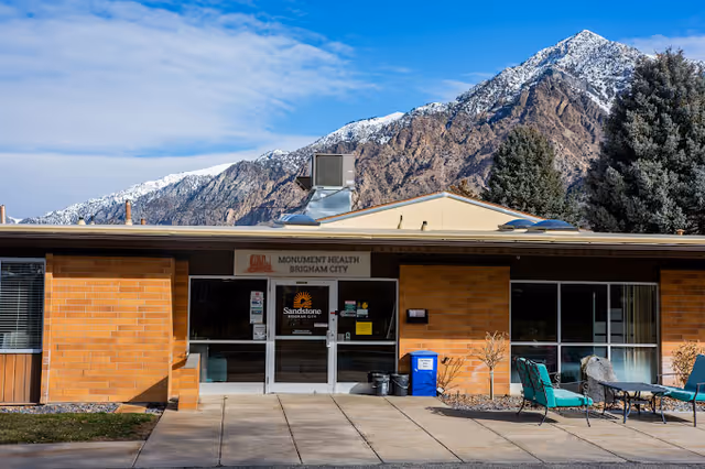Exterior front entrance of a single-story brick building with glass doors, outdoor seating, and snow-capped mountains in the background.