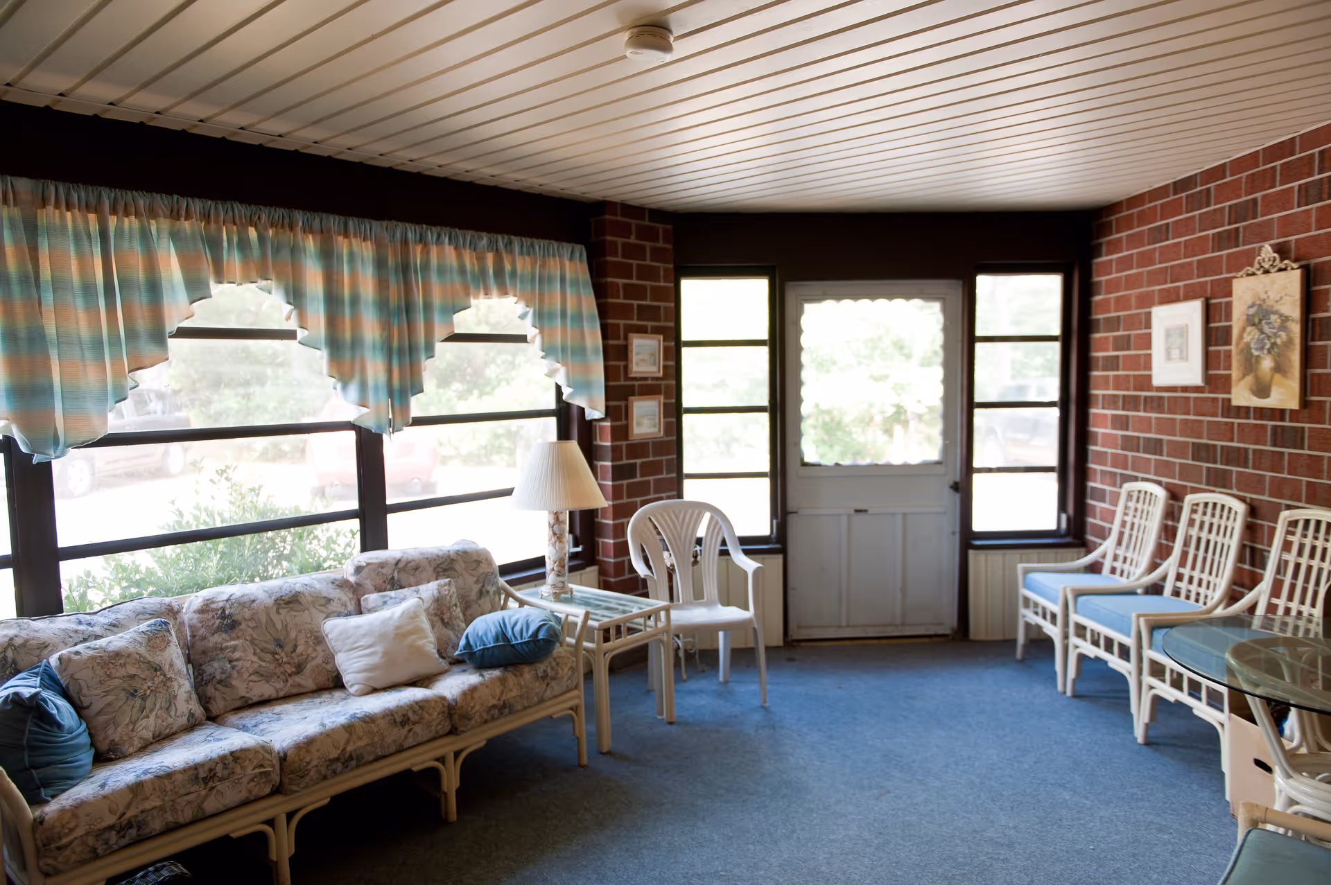 A cozy indoor sitting area with a floral-patterned couch, several white chairs with blue cushions, a small glass-top table, and a lamp. The room has large windows with striped curtains and a door with glass panels, brick walls, and a blue carpeted floor.