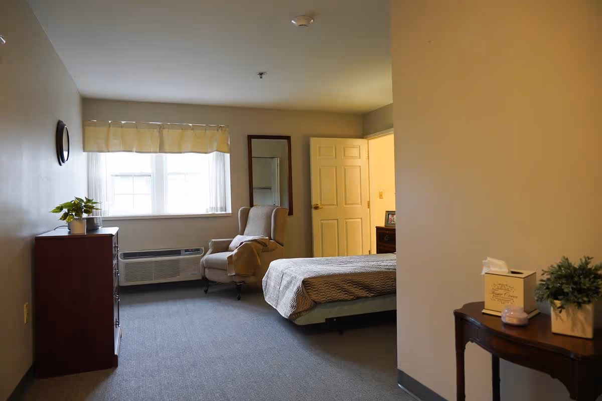 A simple bedroom in a senior living facility with a bed covered in a gray blanket, a beige armchair with a blanket draped over it, a window with light curtains, a dresser with a plant on top, and a small table with a tissue box and a small plant. The door to the room is open, showing a glimpse of another room.