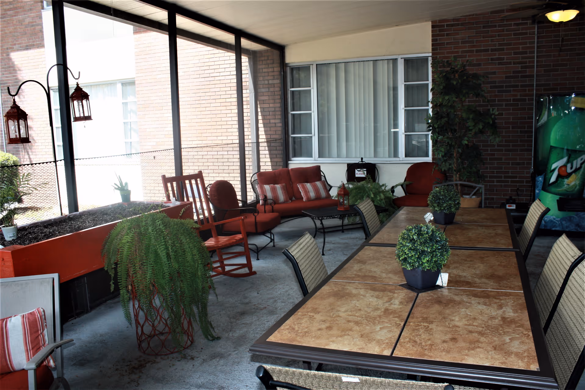 Covered outdoor patio area with a long table and several chairs, potted plants on the table, red cushioned seating including a rocking chair and loveseat, hanging lanterns, and a 7UP vending machine against a brick wall.
