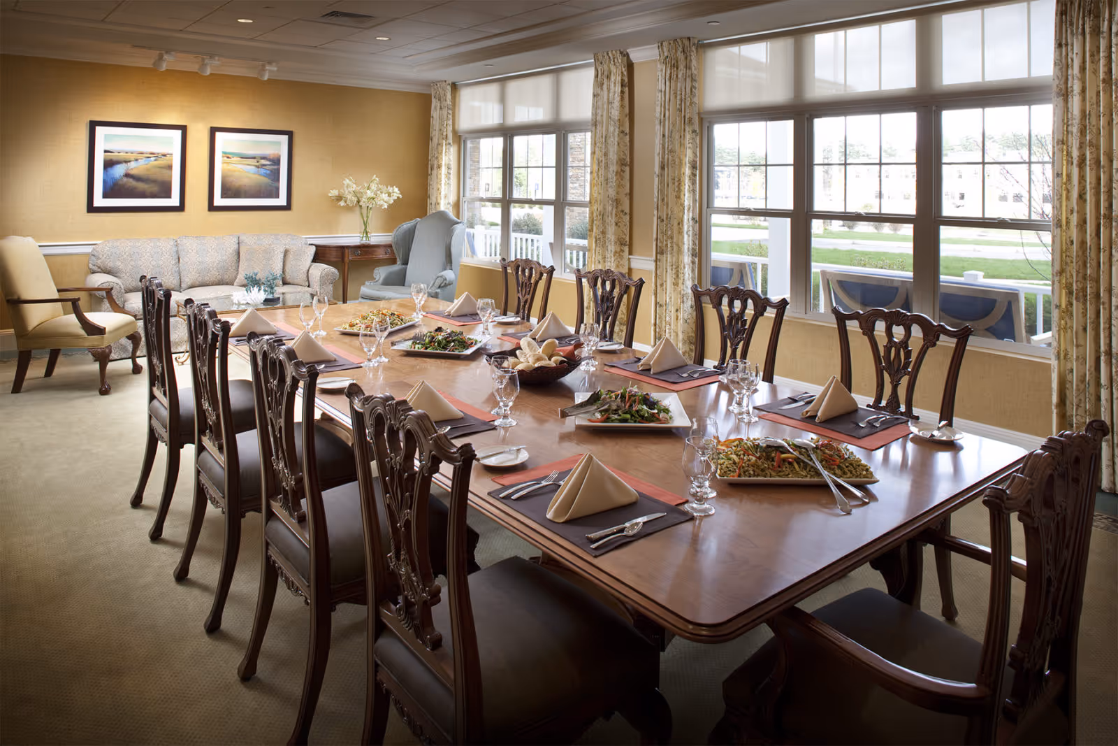 A formal dining room with a large wooden table set for a meal, featuring plates, glasses, napkins, and serving dishes with food. The room has multiple wooden chairs around the table, large windows with patterned curtains letting in natural light, and a seating area with a sofa and armchairs against the wall.