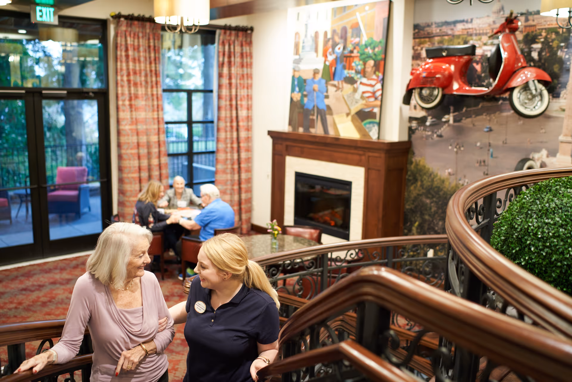An elderly woman and a caregiver are smiling and talking while walking up a curved staircase inside a senior living facility. In the background, three other seniors are seated at a table near large windows with red curtains. The room features a fireplace, colorful artwork on the walls, and a red vintage scooter mounted on the wall as decoration.