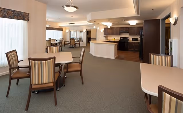 A spacious dining area with several tables and chairs featuring striped upholstery. The room has large windows with sheer curtains allowing natural light to fill the space. In the background, there is an open kitchen with dark wood cabinets, a black refrigerator, stove, and microwave. The flooring transitions from carpet in the dining area to wood in the kitchen. Ceiling lights and wall sconces provide additional lighting.