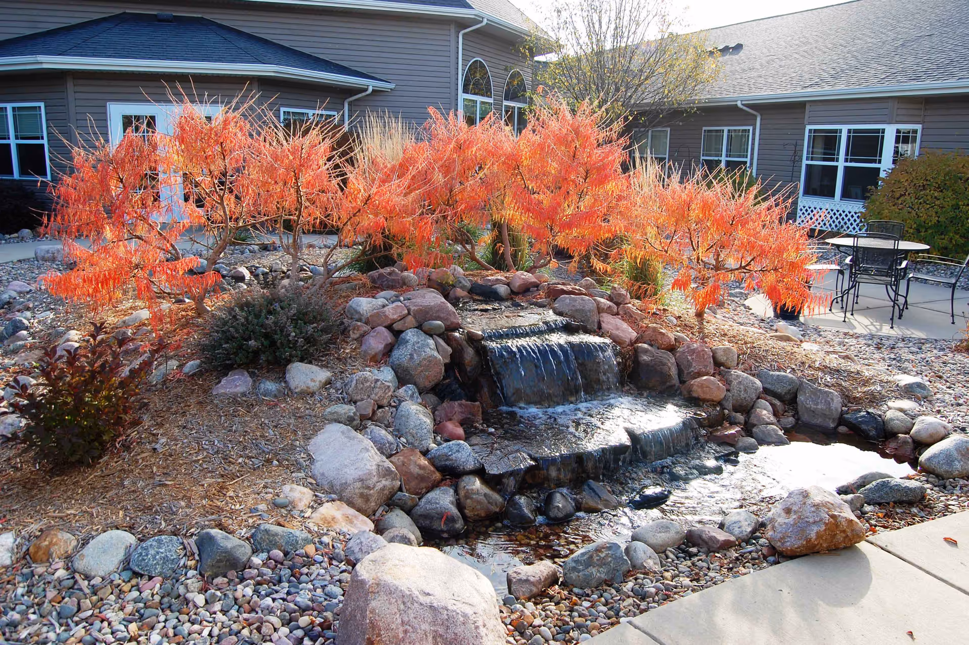 Outdoor garden area at StoneyBrook Suites Assisted Living featuring a small cascading waterfall surrounded by rocks and vibrant orange-red foliage. In the background, there is a patio with metal chairs and tables, and the exterior of the building with windows and doors is visible.
