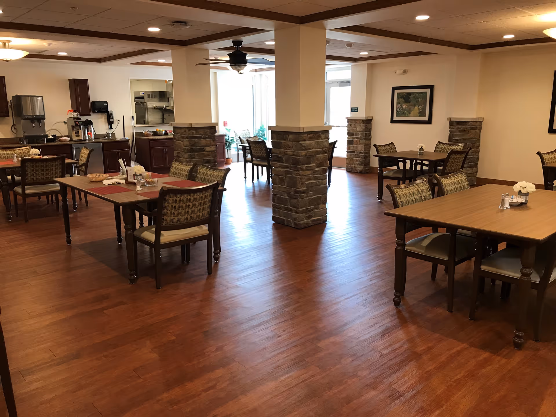 A spacious dining area with wooden floors and several tables and chairs arranged neatly. The room features stone pillars, ceiling fans, and a beverage station with coffee and water dispensers in the background. The walls are decorated with framed pictures, and natural light enters through large windows.