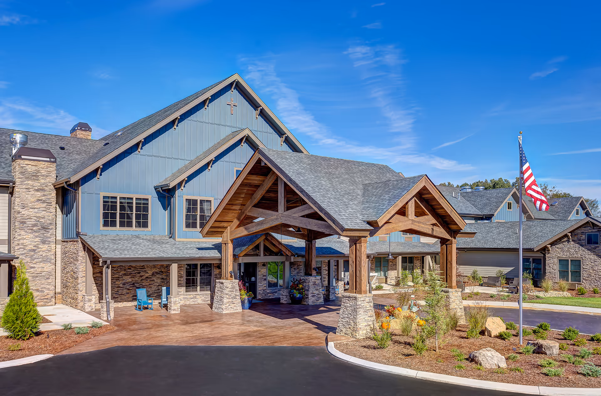 Front entrance and porte-cochère of a large two-story senior living building with wooden beams, stone accents, landscaping, and an American flag.