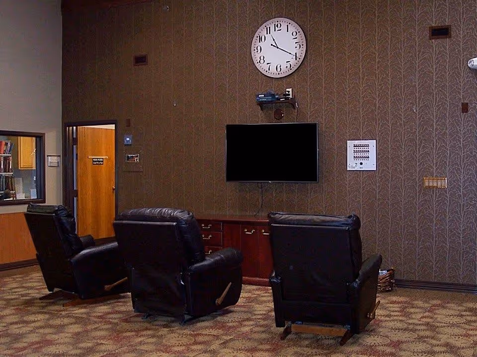 A room with three black leather recliners facing a wall-mounted flat screen TV. Above the TV is a round wall clock showing the time as 11:55. The wall has a brown patterned wallpaper, and below the TV is a wooden cabinet with drawers. To the left, there is a window into another room and a door with a sign. The floor is carpeted with a patterned design.