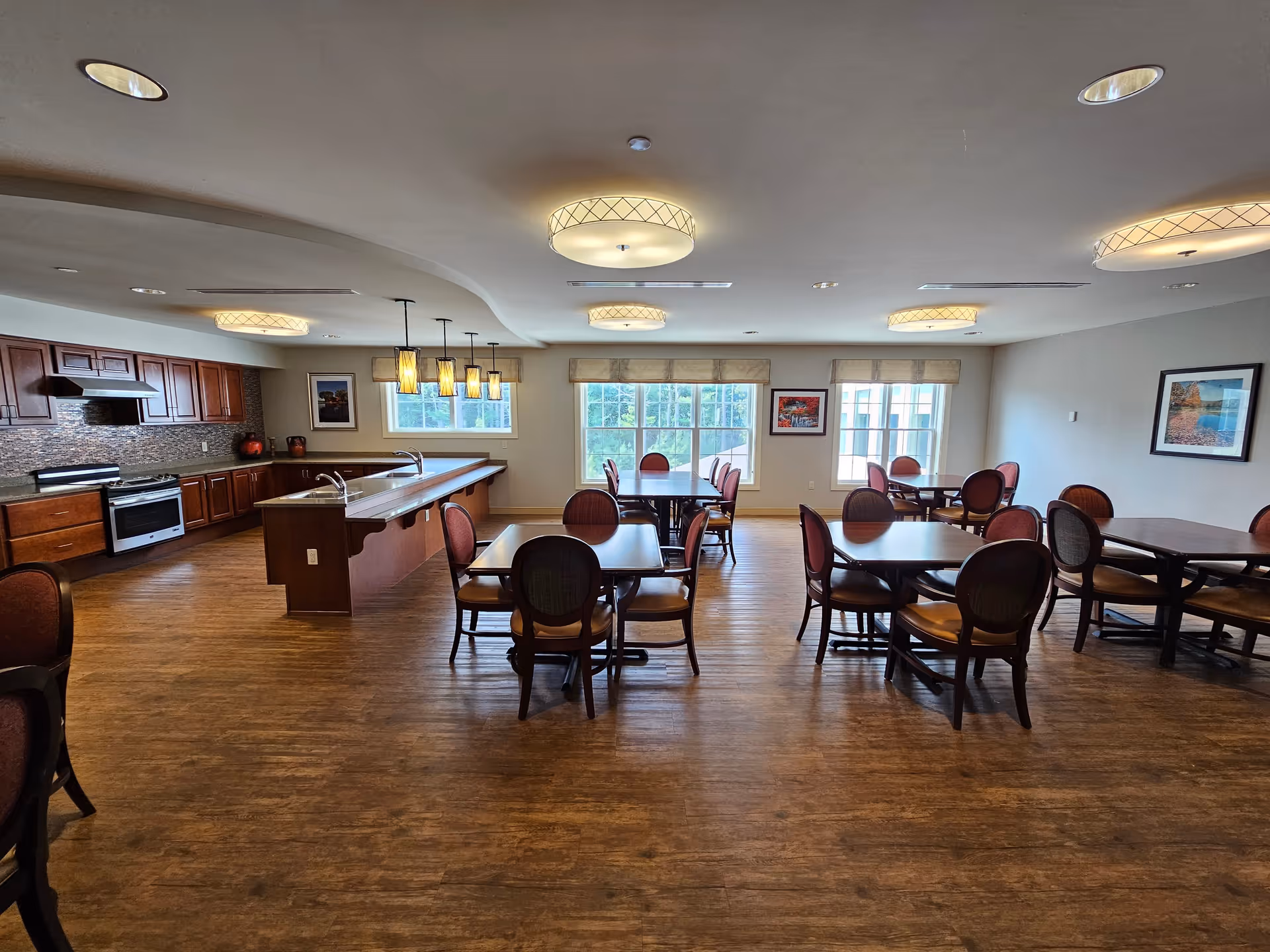 A spacious dining room with multiple tables and chairs arranged neatly. The room features large windows allowing natural light to fill the space, wooden flooring, and a kitchen area with wooden cabinets, a stove, and a long counter with pendant lights hanging above.