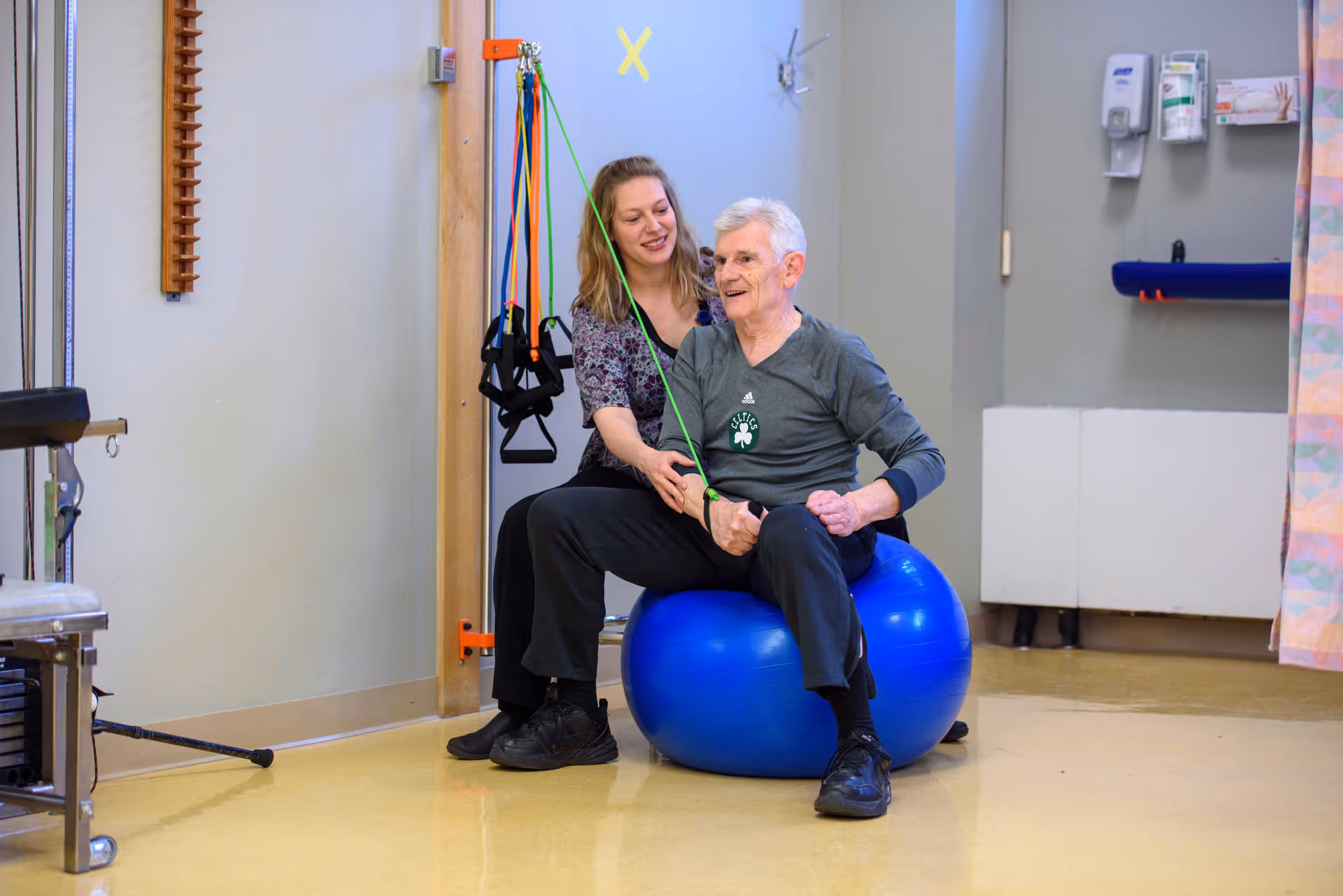 An elderly man sitting on a blue exercise ball while a female therapist assists him with resistance band exercises in a rehabilitation room.
