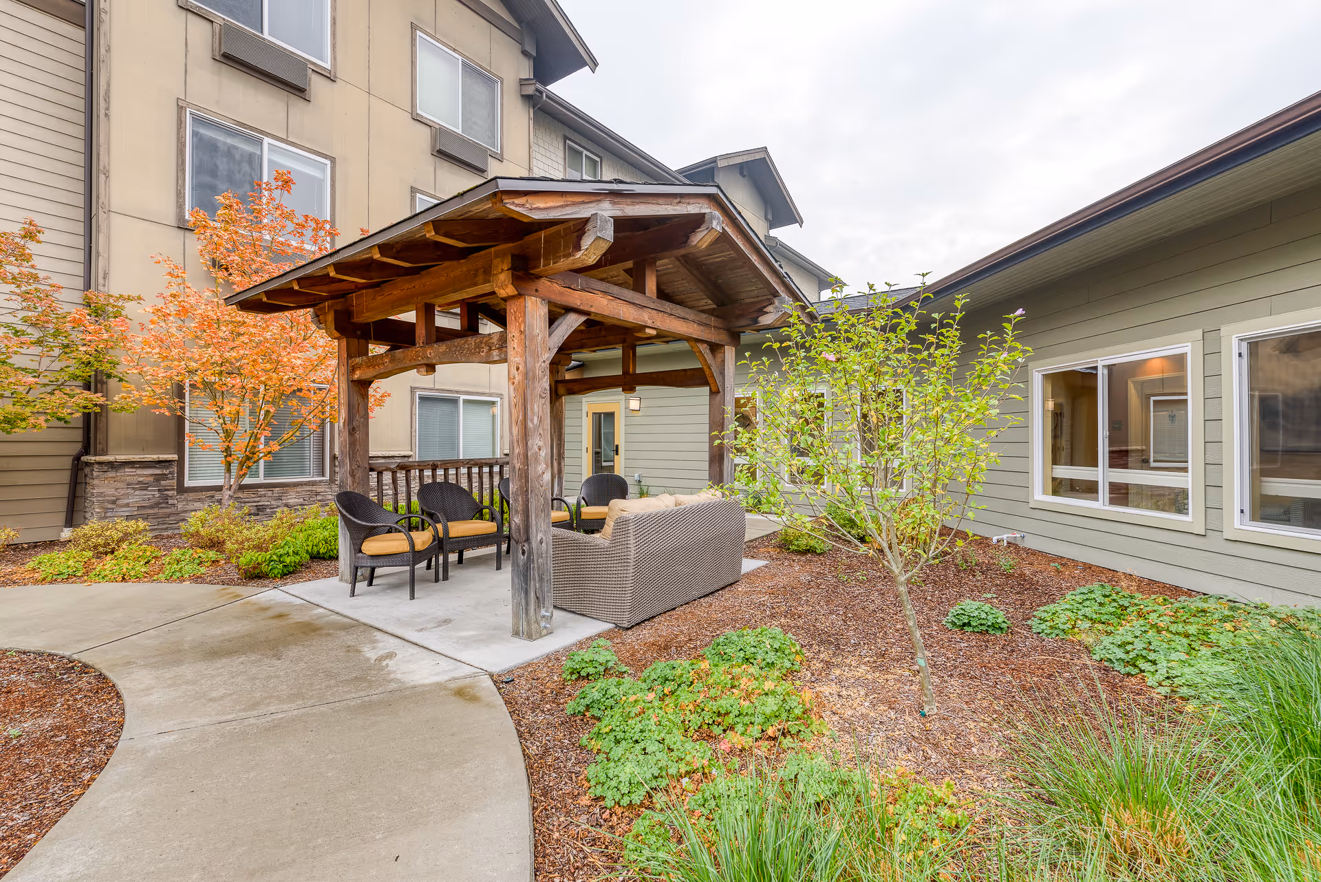 Outdoor seating area at a senior living facility with a wooden pergola covering cushioned chairs and a sofa. The area is surrounded by landscaped plants, small trees, and a curved concrete pathway. The building exterior features beige and green siding with multiple windows.