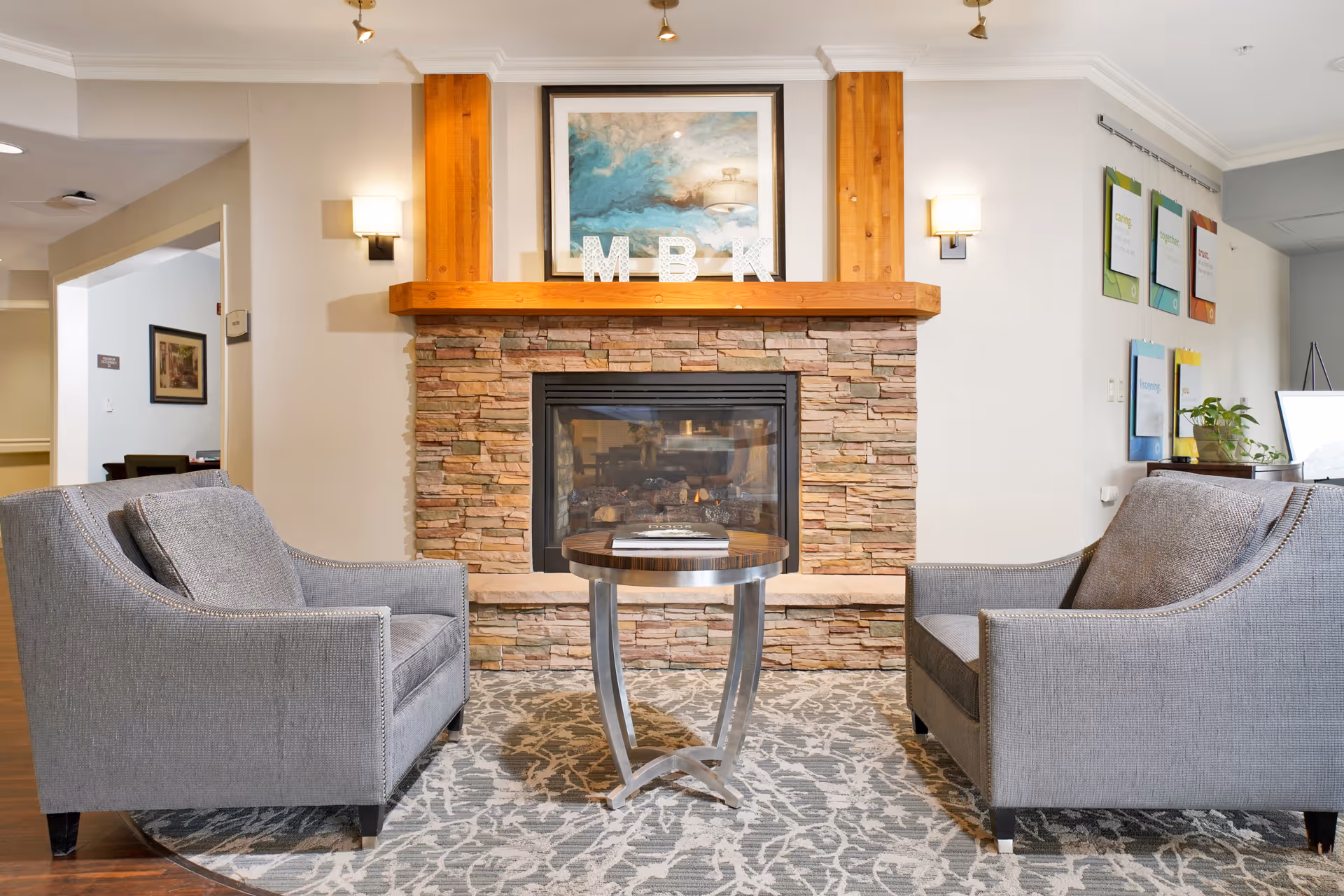 A cozy sitting area with two gray upholstered armchairs facing each other across a small round wooden table, positioned in front of a stone fireplace with a wooden mantel. Above the mantel is a framed abstract painting and decorative letters M, B, and K. The room has soft lighting with wall sconces and a patterned area rug on the floor.