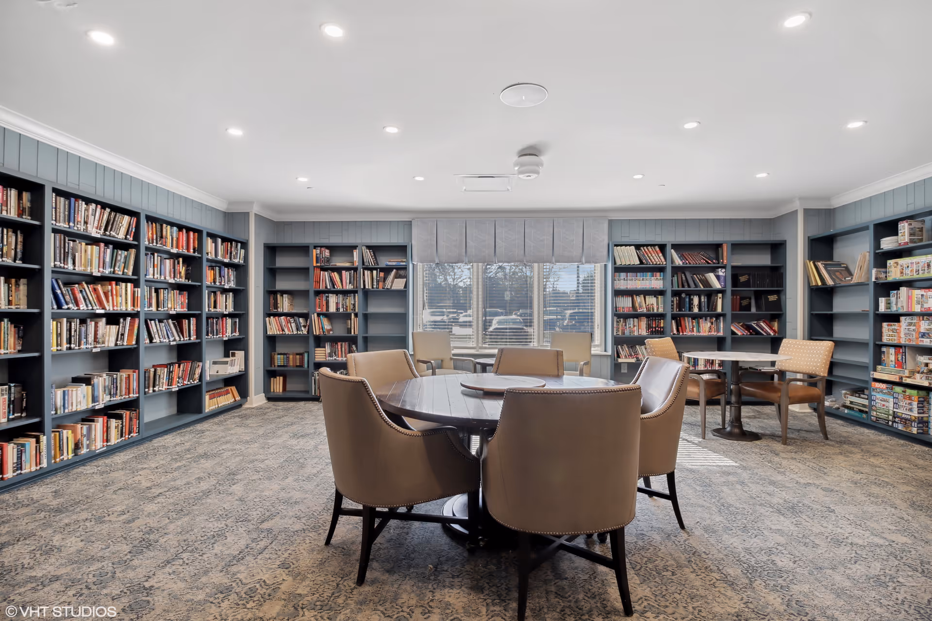A bright and spacious library room with multiple bookshelves filled with books along the walls. In the center, there is a round wooden table surrounded by six beige upholstered chairs. Additional seating with chairs and small tables is placed near a large window that lets in natural light. The room has a carpeted floor and recessed ceiling lights.