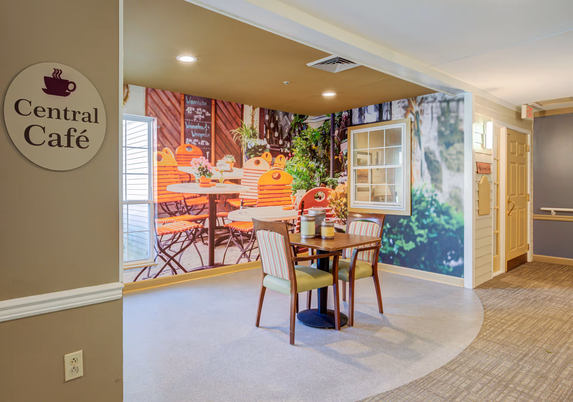 Indoor seating area with a small wooden table and two chairs with striped cushions. The wall behind features a large mural of an outdoor café scene with orange chairs and tables, plants, and a chalkboard menu. A round sign on the left wall reads 'Central Café'. The floor is a mix of carpet and smooth flooring, and there is a hallway with doors on the right side.