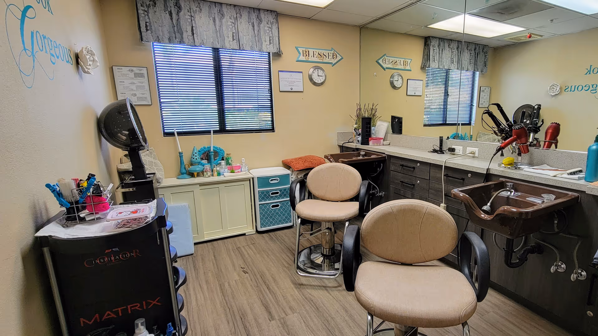 Interior salon room with two styling chairs, shampoo sinks, mirrors, and cabinets in a senior living facility.
