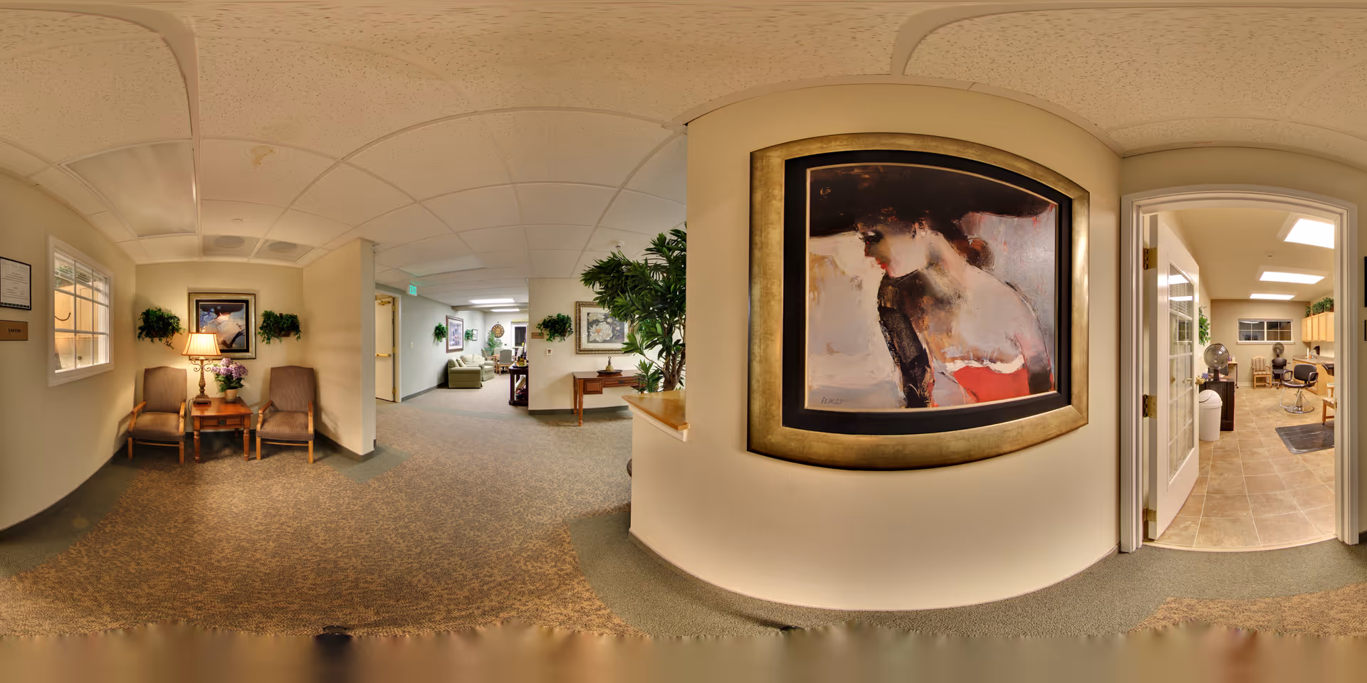 Interior view of a senior living facility hallway with beige walls and carpeted floor. There are two chairs with a small table and lamp between them on the left side, decorated with a framed painting and plants. Further down the hallway, there is a seating area with sofas and more artwork on the walls. On the right side, an open doorway leads to a room with tile flooring, salon chairs, and hair dryers, suggesting a beauty or barber area.