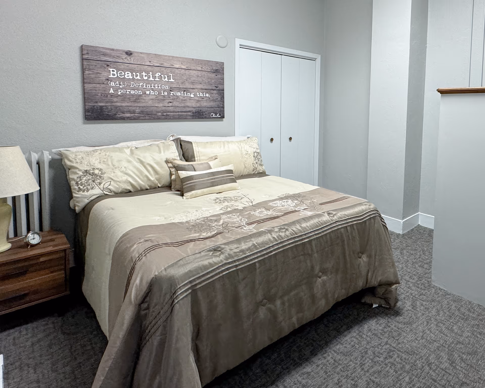 Bedroom with a made bed, decorative pillows, a wooden nightstand with lamp, and a framed sign reading "Beautiful" on the wall.