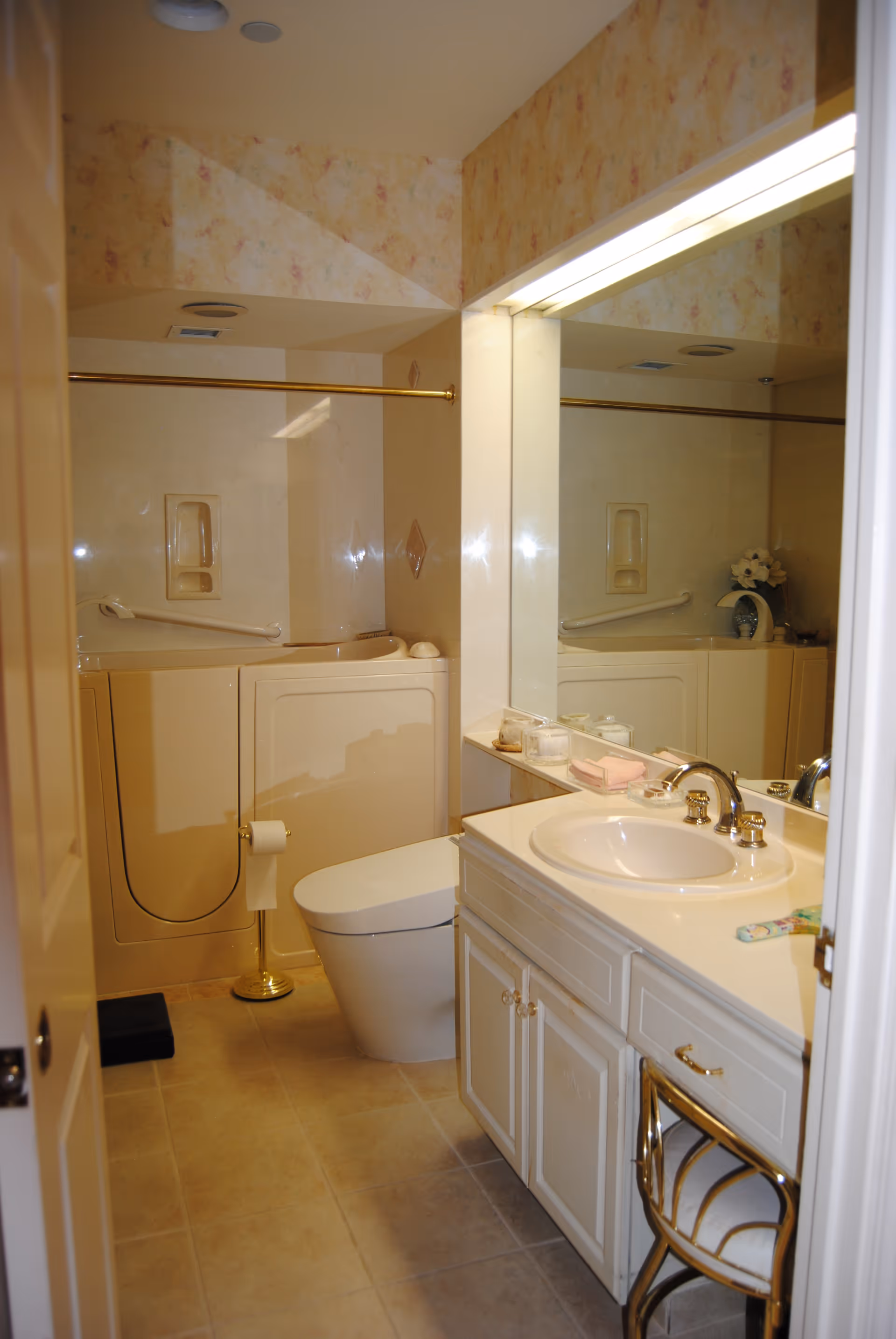 A bathroom with a walk-in bathtub, a toilet, and a sink with a countertop. The walls have a light floral wallpaper, and there is a large mirror above the sink. A gold-colored towel bar is mounted above the bathtub, and a small chair with a gold frame is placed under the sink counter.