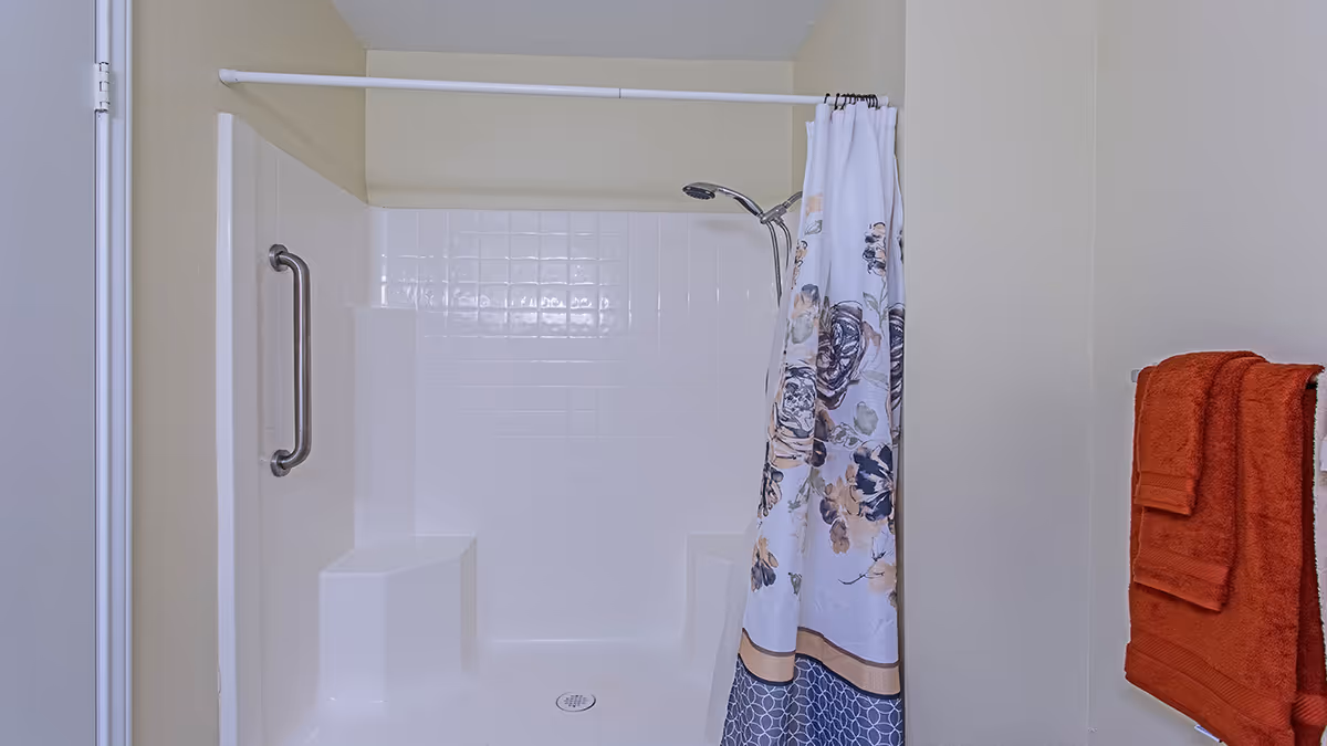 White tiled shower stall with built-in seats, a stainless grab bar, floral shower curtain, and rust-colored towels on a wall rack.