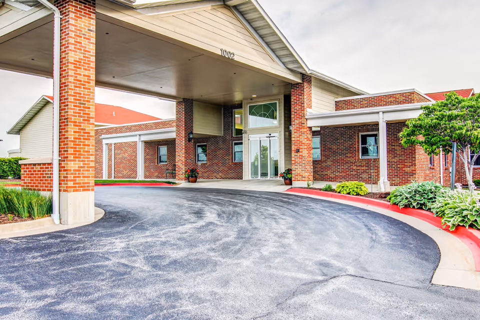Covered porte-cochere and curved driveway leading to glass double doors at the brick entrance of a senior living building.