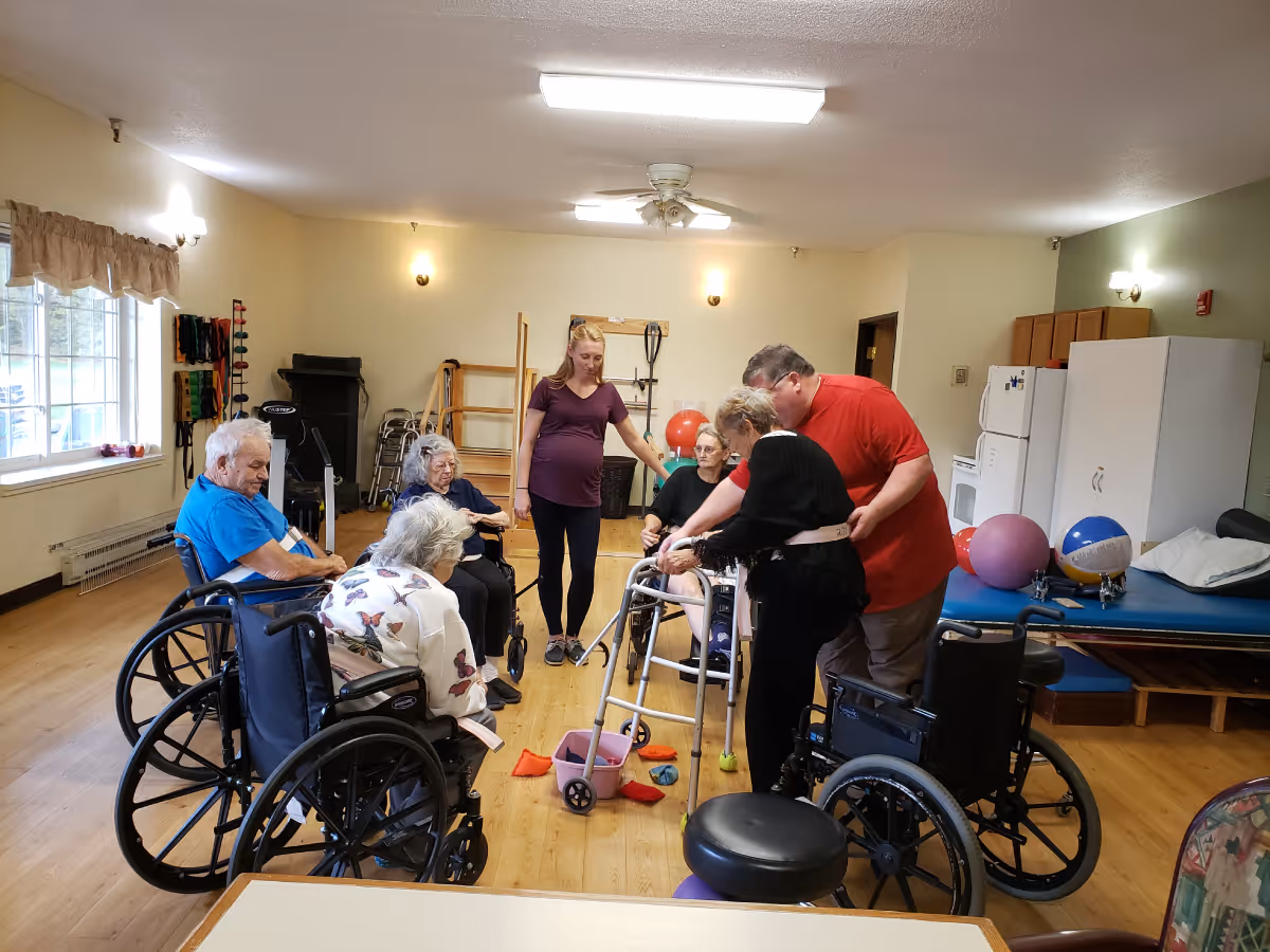 A group of elderly people, some in wheelchairs, participating in a physical therapy or exercise session in a well-lit room with wooden floors. A young woman and a man are assisting the elderly individuals, one of whom is using a walker. The room contains exercise equipment, therapy balls, and a refrigerator in the background.