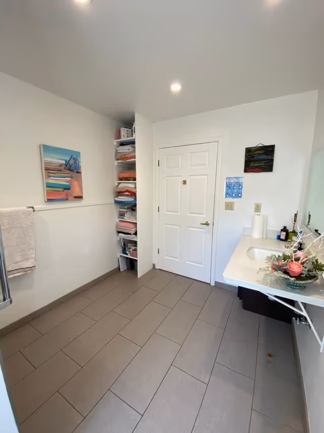 A clean and spacious bathroom with beige tiled floor, white walls, and a white door. On the left wall, there is a towel rack with a white towel and a colorful painting above it. Next to the door, there is a built-in shelving unit filled with neatly folded towels and other items. On the right side, there is a white countertop with a sink, a floral arrangement, paper towels, and some toiletries. The ceiling has recessed lighting.