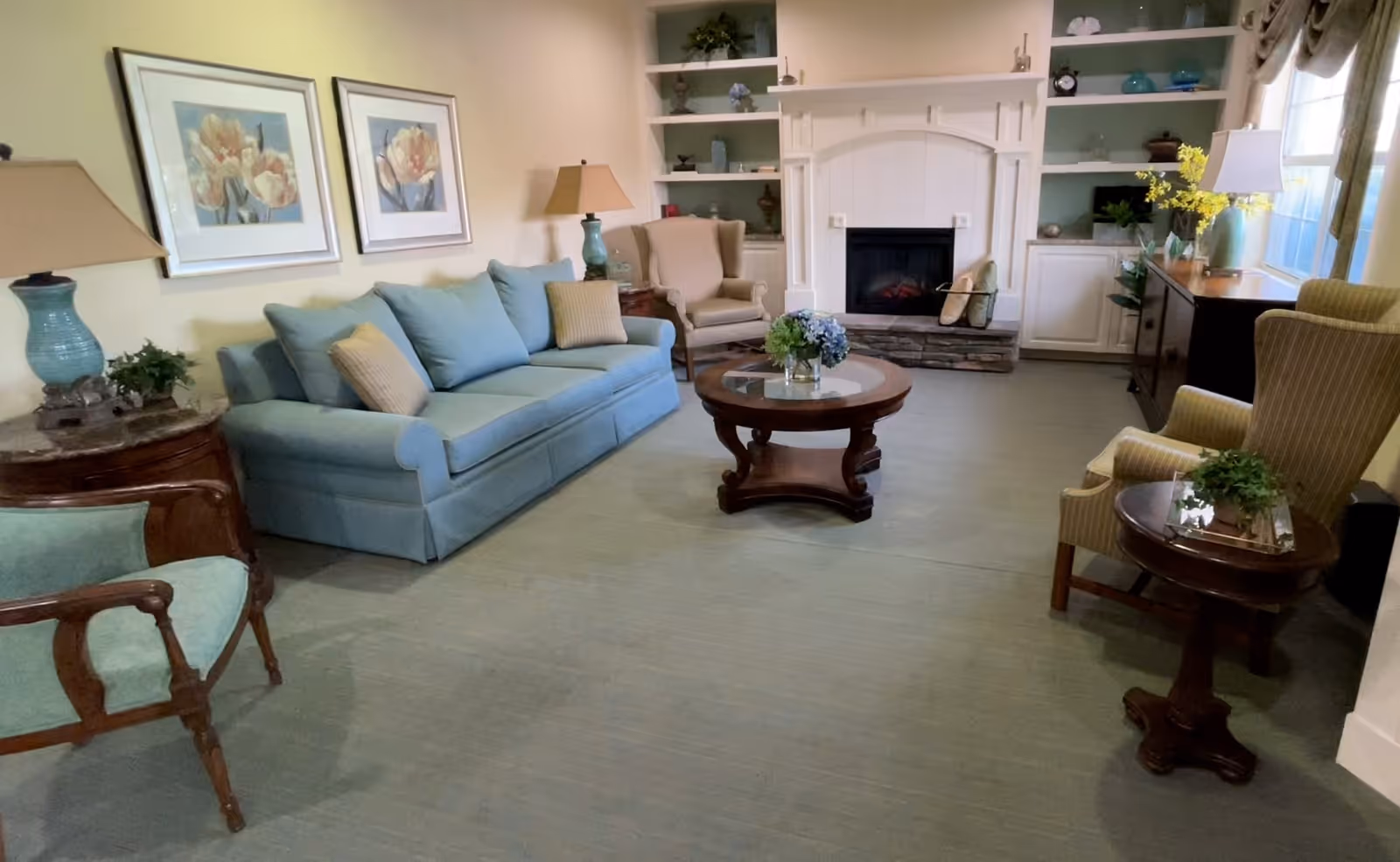 A cozy living room with a light blue sofa, two armchairs, a round wooden coffee table with a flower arrangement, built-in shelves flanking a white fireplace, two framed floral paintings on the wall, and table lamps on side tables. The room has a soft, neutral color palette with natural light coming through a window.