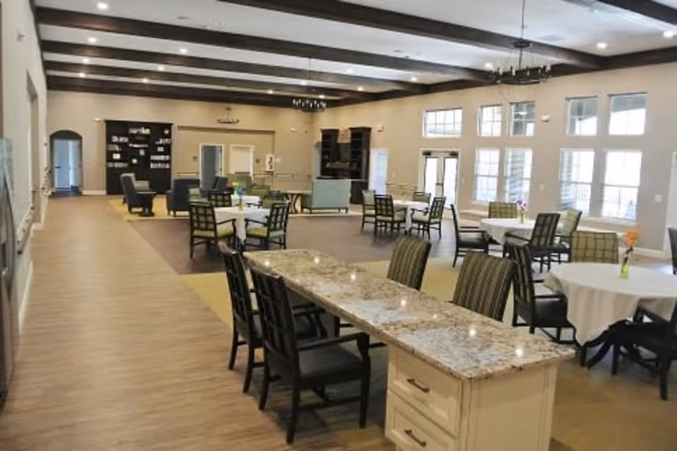 Spacious senior living facility common area with multiple round tables covered with white tablecloths and chairs arranged around them. A long granite countertop island with chairs is in the foreground. The room has large windows letting in natural light, wooden ceiling beams, and comfortable seating areas with armchairs and sofas in the background.