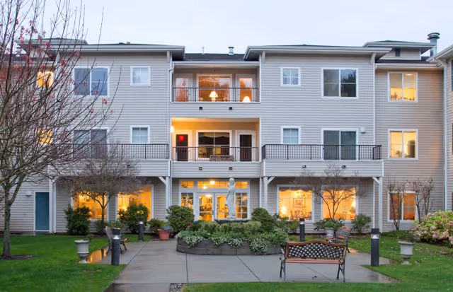 Exterior view of a three-story senior living facility building with multiple windows and balconies. The foreground features a paved walkway, green lawn, landscaped bushes, small trees, and a bench. The building is light-colored with outdoor lighting visible through the windows.