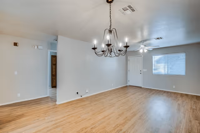 Empty room with light wood flooring, beige walls, a black chandelier hanging from the ceiling, a ceiling fan with light, a white door, and a window with blinds.