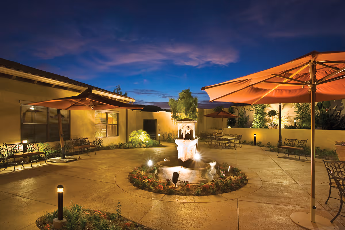A lit outdoor courtyard at dusk featuring a central fountain surrounded by flower beds, patio umbrellas, benches, and seating.