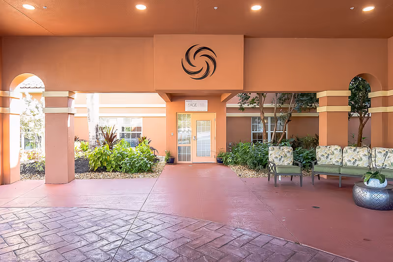 Covered entryway of a senior living facility with seating, potted plants, and a glass door under an orange stucco archway.