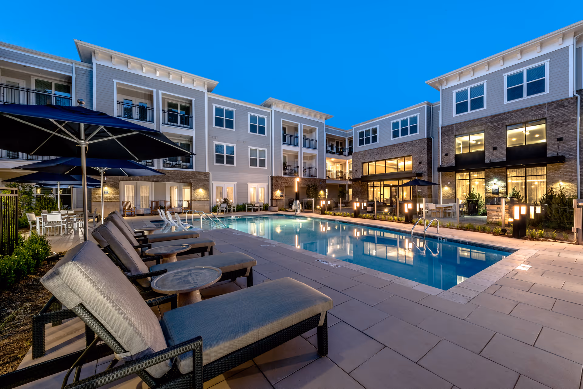 Outdoor courtyard with a lit swimming pool, lounge chairs, umbrellas, and a multi-story residential building at dusk.