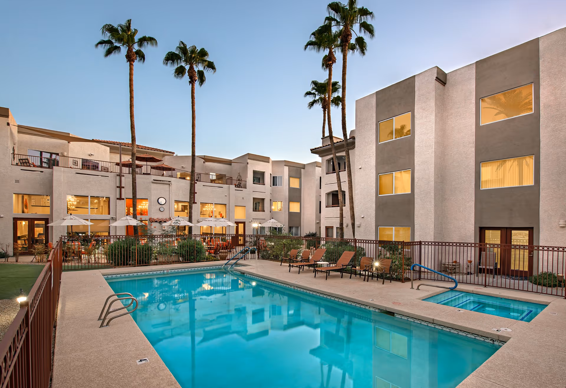 Multi-story senior living building surrounding a courtyard with a swimming pool, lounge chairs, umbrellas, and tall palm trees at dusk.