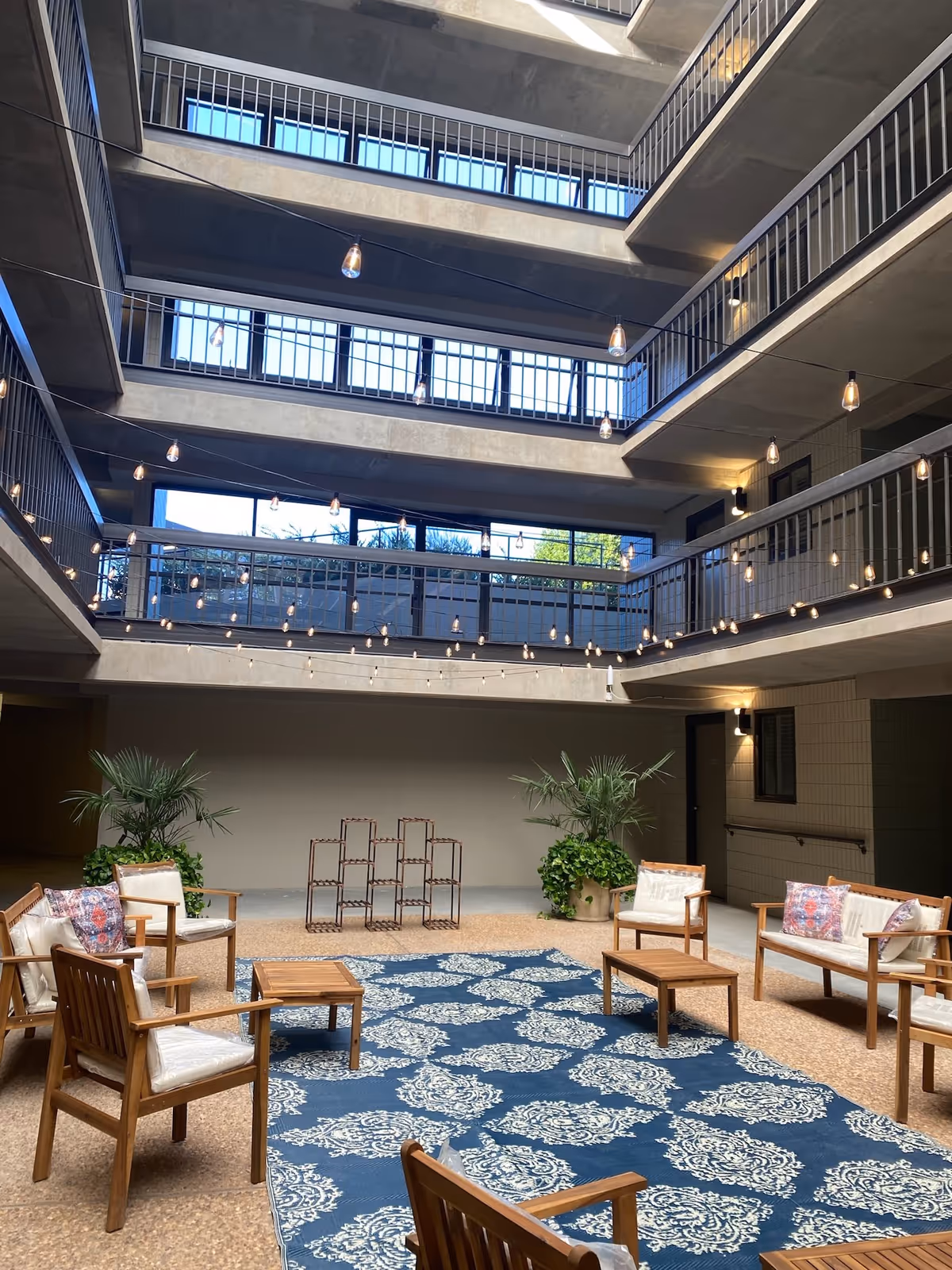Indoor courtyard area with multiple floors visible, featuring string lights hanging across the open space. The ground level has wooden chairs and sofas with cushions arranged around a blue patterned rug, along with potted plants and a decorative metal rack against the wall.
