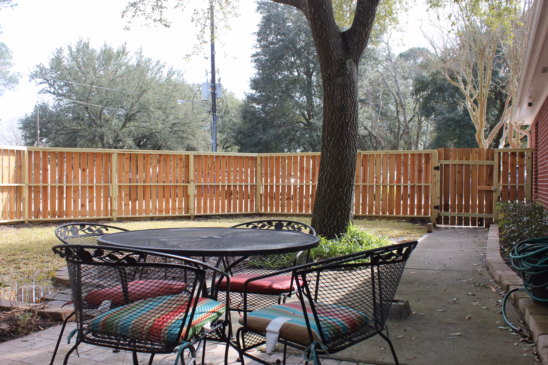 Outdoor patio area with a round metal table and four metal chairs with colorful cushions. The patio is surrounded by a wooden fence, and there is a large tree near the table. A garden hose is coiled on the right side near a brick building wall.