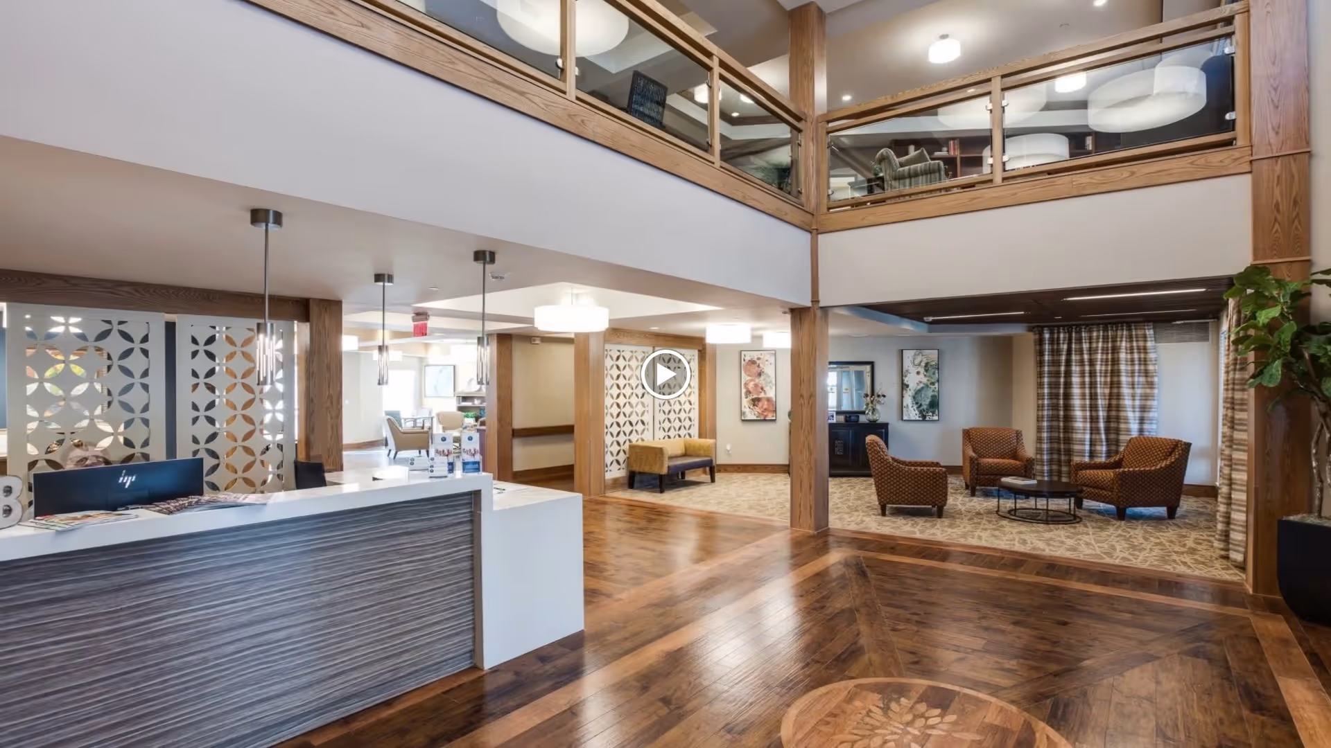 Spacious and well-lit senior living facility lobby with a modern reception desk on the left, wooden flooring with a decorative inlay, seating area with four armchairs and a round table on the right, and a second-floor balcony with glass railing overlooking the lobby.