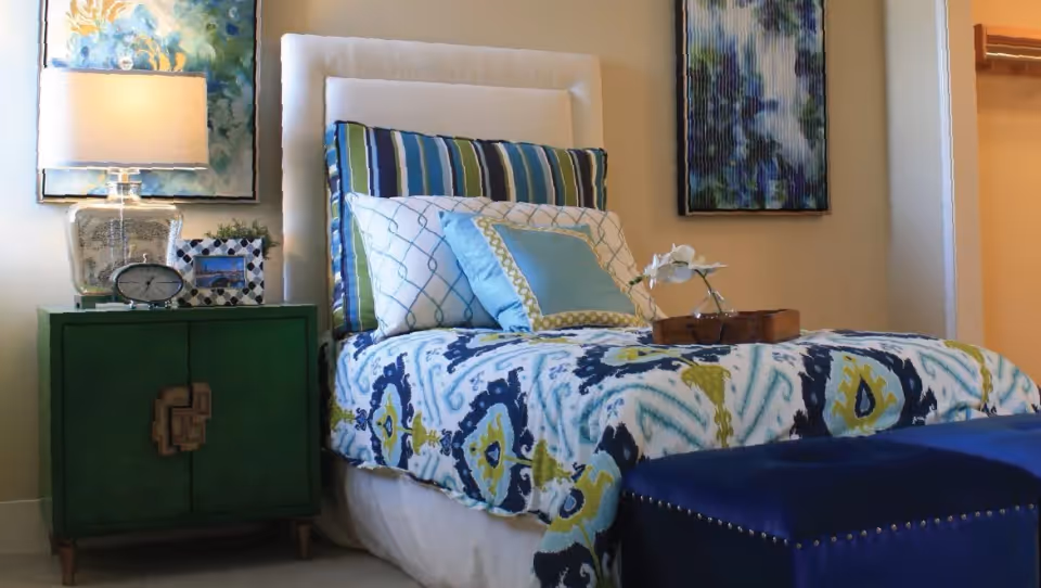 A neatly made bed with a white upholstered headboard, patterned blue, green, and white bedding, and multiple pillows. Next to the bed is a green nightstand with a decorative lamp, a clock, and a framed photo. Two abstract paintings hang on the wall above the bed and nightstand. A blue ottoman is placed at the foot of the bed.