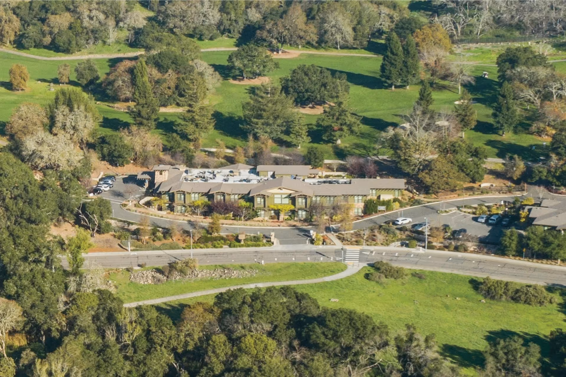 Aerial view of a single-story senior living building with surrounding parking, roads, and green lawns and trees.