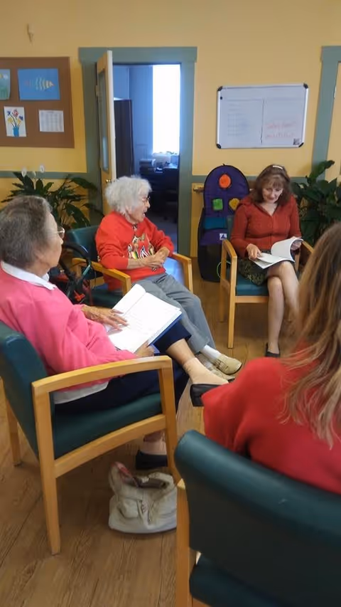 A group of elderly women sitting in a circle in a room, engaged in reading or discussion. The room has yellow walls with blue trim, a bulletin board with artwork, a whiteboard, and some plants. The floor is wooden, and the women are seated on green cushioned chairs with wooden armrests.