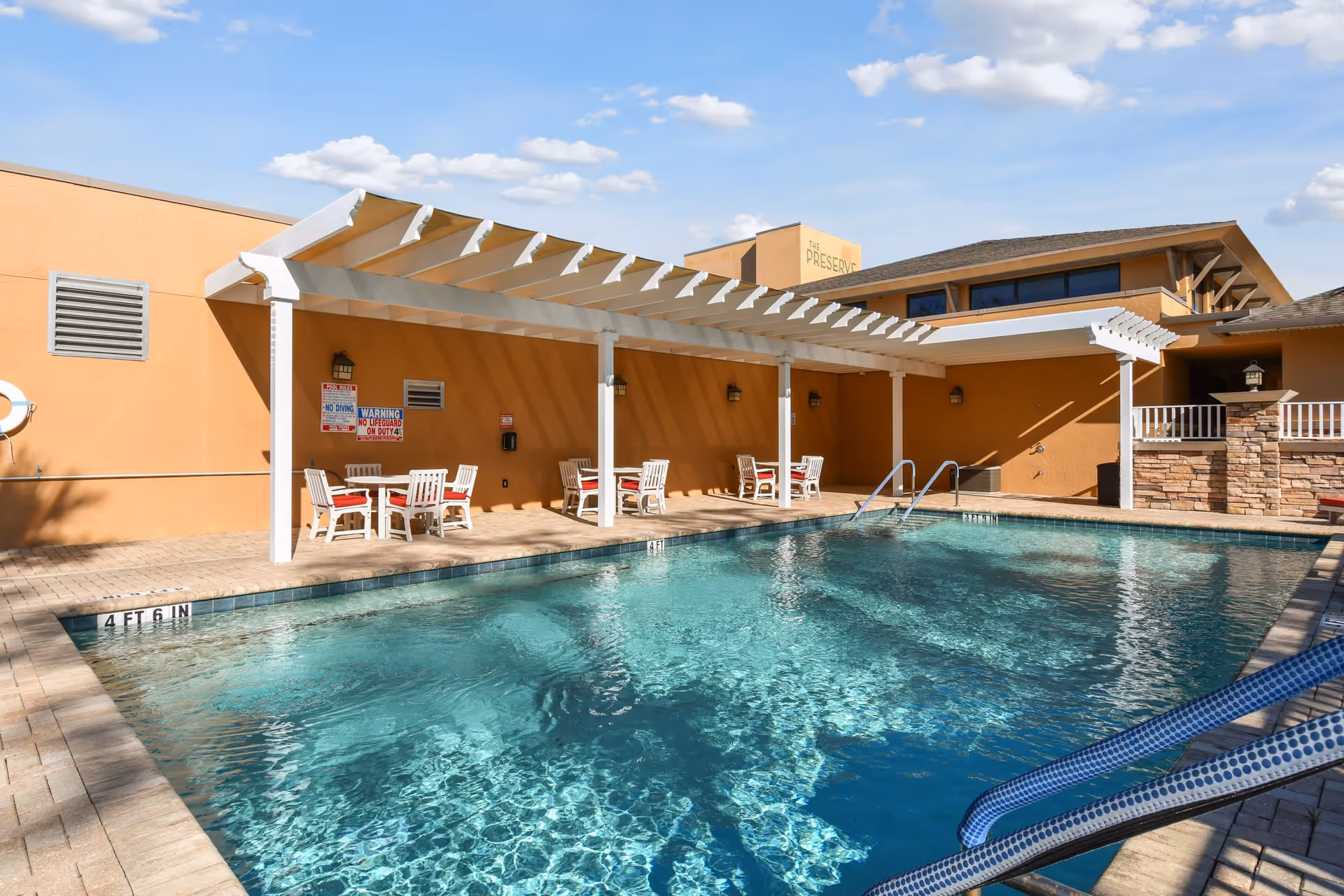 Outdoor swimming pool area at The Preserve at Dunedin with clear blue water, surrounded by a beige tiled deck. There is a white pergola providing shade over several white tables and chairs with red cushions. The building in the background is painted in a warm beige color with a sign that reads 'The Preserve'. The sky is blue with scattered clouds.