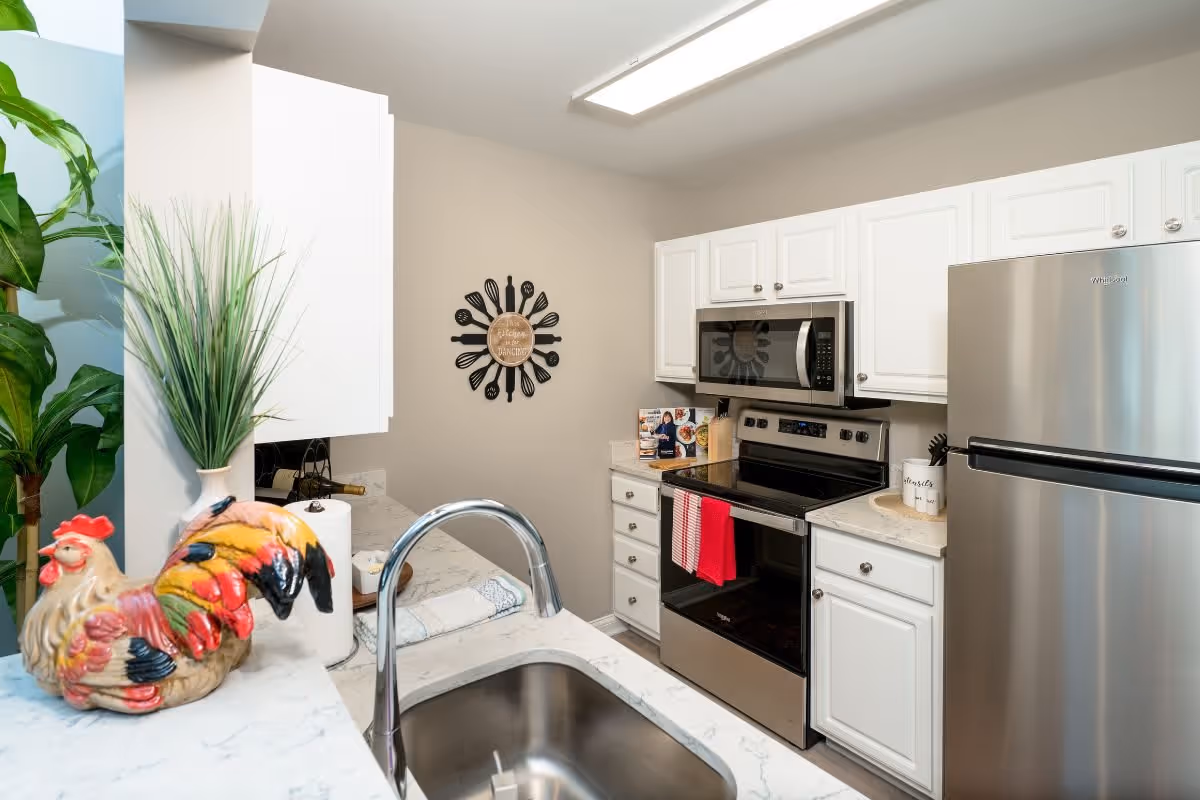 Modern kitchen with white cabinets, stainless steel refrigerator and oven, a sink with a chrome faucet, a decorative ceramic rooster, a potted plant, and a wall decoration featuring kitchen utensils arranged in a circular pattern.
