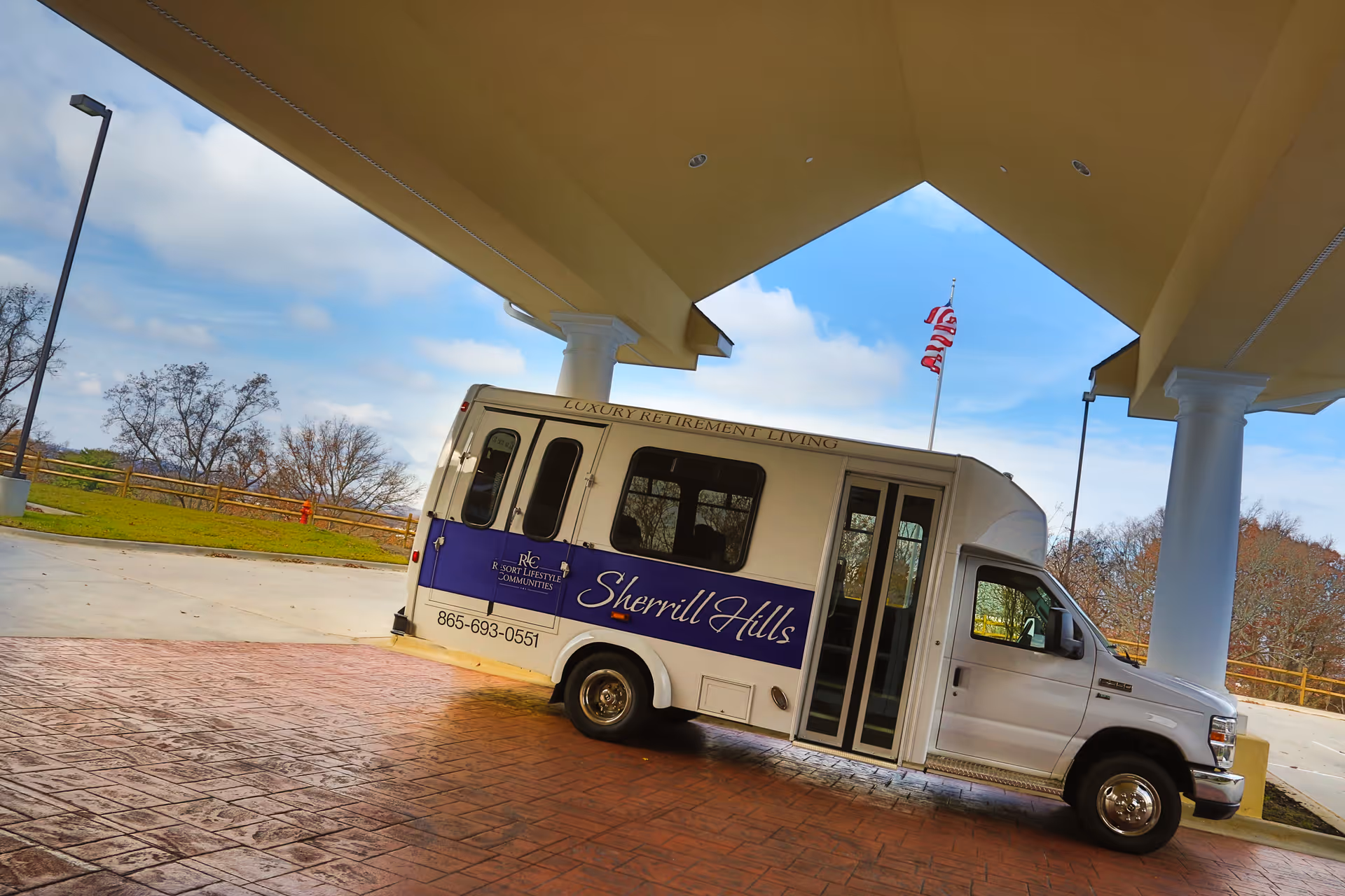A white shuttle bus with purple signage parked under a covered entrance. The bus has the text 'Sherrill Hills' and 'Luxury Retirement Living' on its side along with a phone number. An American flag is visible in the background against a partly cloudy sky.