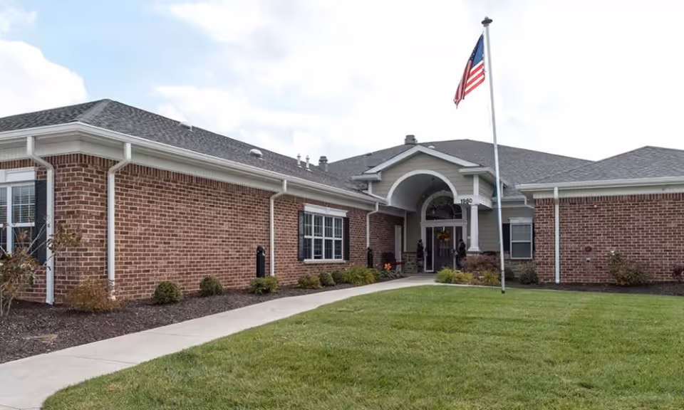 Exterior view of a single-story brick building with a gray shingled roof, a curved concrete walkway leading to the main entrance, and an American flag on a flagpole in front. The entrance features a covered archway with white columns, and there are small shrubs and landscaping along the building's foundation.